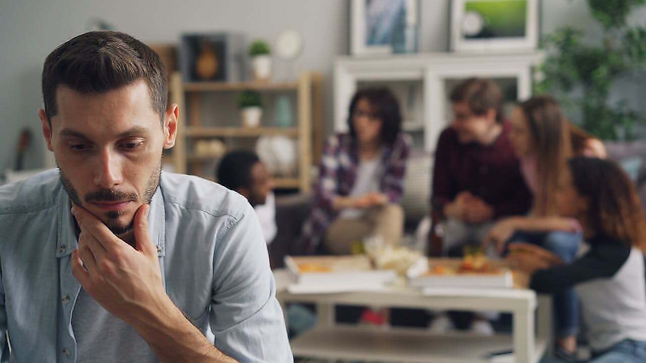 A man in a living room clearly being an outcast of others at a table behind him.