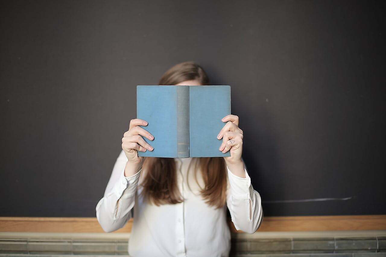 The Humbling Process in Becoming a True Teacher. Humility has shown up for me again and again and not kindly I must say. Image of a woman standing in front of a chalk board holding a book in front of her face.